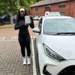 Barbara holding her driving test pass certificate next to Khan School of Motoring car in Slough